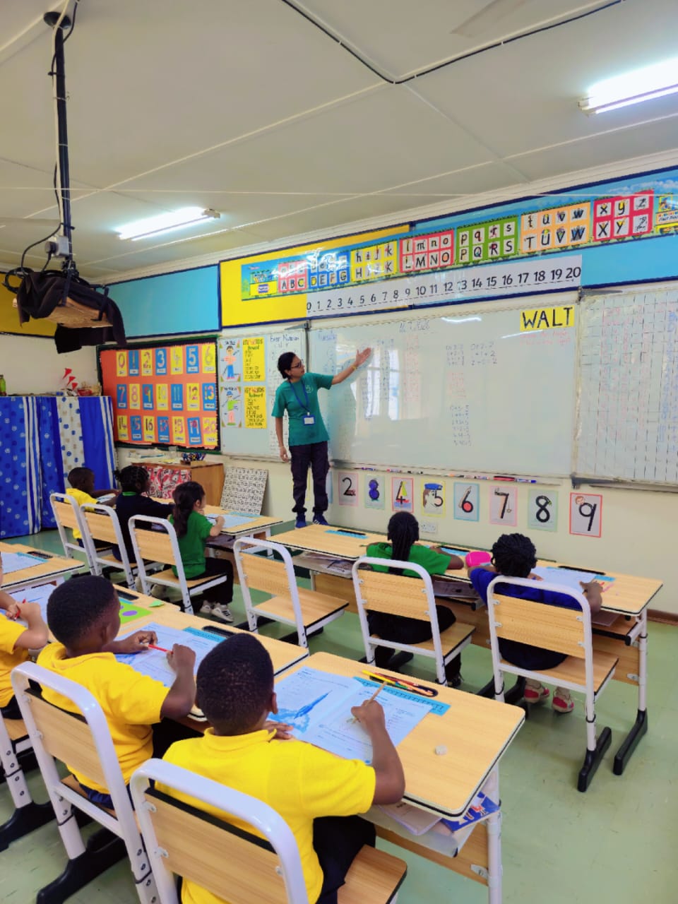 A teacher leads number work at the whiteboard while the class follows along in their books.